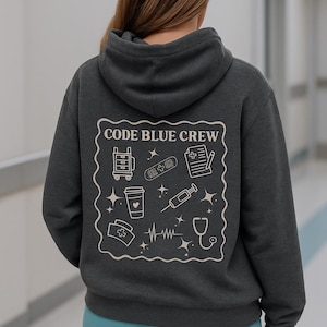 Puede incluir: Sudadera con capucha gris oscuro con un diseño en la espalda que dice "CODE BLUE CREW" con ilustraciones médicas. El diseño incluye un carro médico, una taza de café, una venda, una jeringa y un estetoscopio.