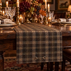 Puede incluir: Un camino de mesa rectangular con un patrón de cuadros en marrón oscuro y beige. El camino de tela está colocado sobre una mesa de madera y presenta un detalle de herraje metálico dorado en forma de bocado de caballo cerca del borde inferior.