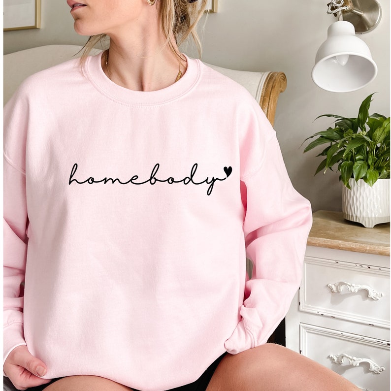 May include: Pink sweatshirt with the text "homebody" in black cursive lettering and a small black heart.