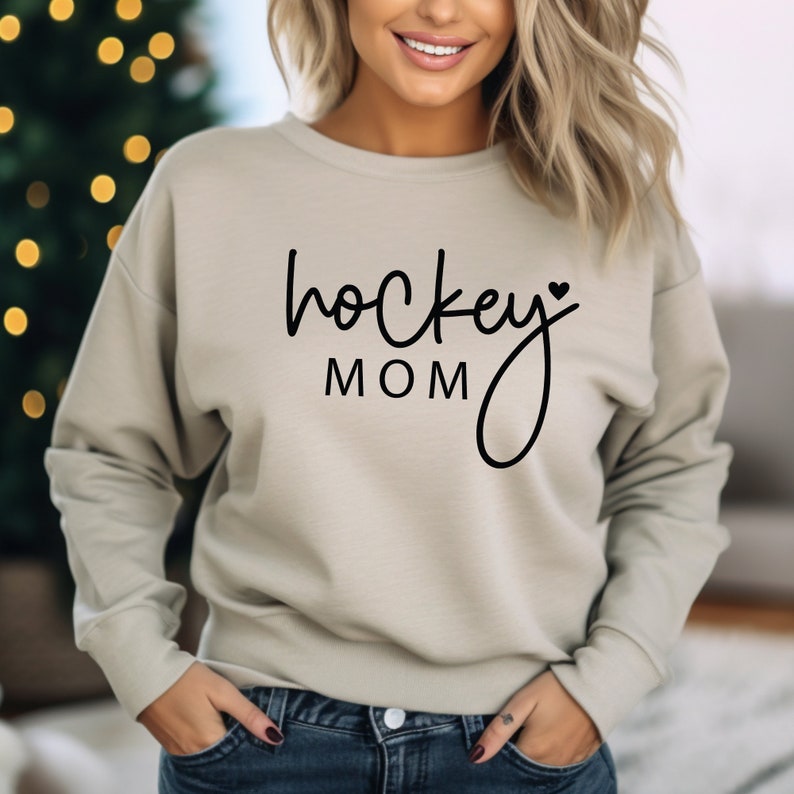 Pu&ograve; includere: Una felpa beige chiaro con la scritta "hockey mom" in nero con un piccolo simbolo di cuore.