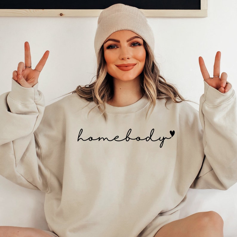 May include: A light beige sweatshirt with the text "homebody" in black script with a small heart at the end. The model is wearing the sweatshirt and a light beige beanie.