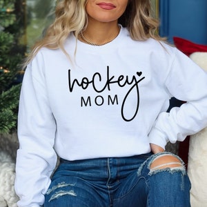 Pu&ograve; includere: Felpa bianca con testo nero che dice "hockey mom" con un piccolo cuore dopo la parola "hockey".