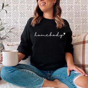 May include: A woman wearing a black sweatshirt with the text "homebody" in white with a small heart symbol. She is sitting on a white surface with a brown and white plaid throw pillow behind her.