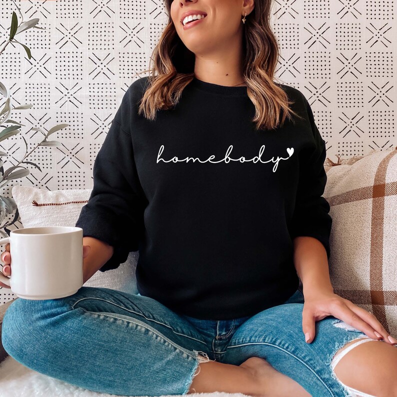 May include: A woman wearing a black sweatshirt with the text "homebody" in white with a small heart symbol. She is sitting on a white surface with a brown and white plaid throw pillow behind her.