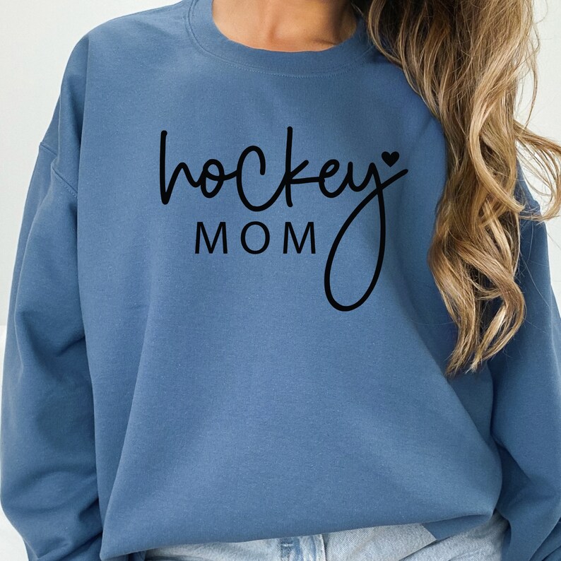 Pu&ograve; includere: Una felpa blu con il testo "hockey mom" in corsivo nero con un piccolo simbolo di cuore.