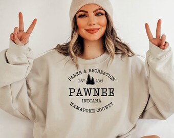 Peut inclure: Un sweat-shirt beige avec un imprimé graphique noir qui dit "Parks & Recreation Est. 1817 Pawnee Indiana Wapapoke County".