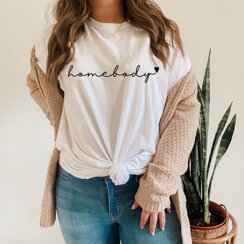May include: White t-shirt with the text "homebody" in black cursive lettering with a small black heart at the end. The shirt is tied at the front and worn with blue jeans and a beige cardigan.