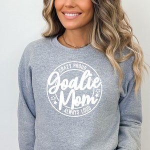 K&ouml;nnte beinhalten: Grauer Pullover mit einem wei&szlig;en Kreis-Motiv, auf dem "Crazy Proud Goalie Mom Always Loud" steht.