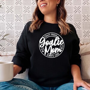 K&ouml;nnte beinhalten: Schwarzer Pullover mit einem wei&szlig;en Kreis-Motiv, auf dem "Crazy Proud Goalie Mom Always Loud" steht.