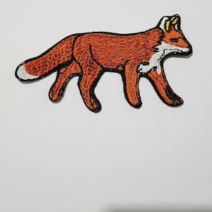 Puede incluir: Parche bordado con un zorro caminando. El zorro es principalmente naranja con detalles blancos y negros. El parche tiene un contorno negro. La cola del zorro tiene una punta blanca.