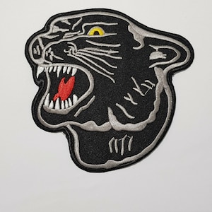 May include: Embroidered patch of a roaring black panther head with a grey outline. The panther's mouth is open, showing white teeth and a red tongue. The eyes are yellow.
