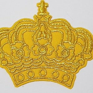 Puede incluir: Un parche bordado en oro con forma de corona. La corona está detallada con intrincados bordados y tiene una punta puntiaguda.