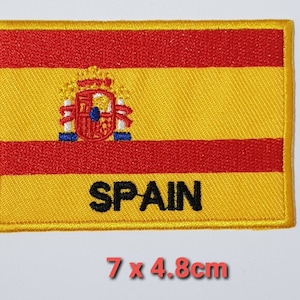 May include: Embroidered patch of the Spanish flag with the coat of arms in the center and the word "SPAIN" below. The patch is yellow, red, and has a yellow border. It measures 7 by 4.8 centimeters.