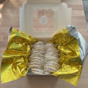 May include: An open white bakery box with a clear window, filled with individually wrapped, round, powdered sugar-dusted cookies. The cookies are nestled in gold and silver foil. The box sits on a light brown wooden surface.