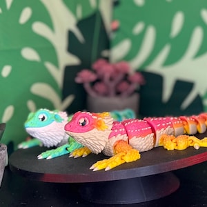 Peut inclure: Deux figurines de lézard articulées, roses, oranges, jaunes et turquoise, reposent sur une plateforme noire. Le fond présente un motif de feuilles tropicales vertes et des plantes succulentes en pot. Les figurines mesurent environ 20 cm de long.