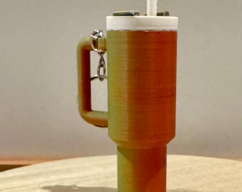May include: A 3D printed keychain shaped like a cup of coffee with a straw. The keychain is orange and yellow with a white lid and a silver metal ring.