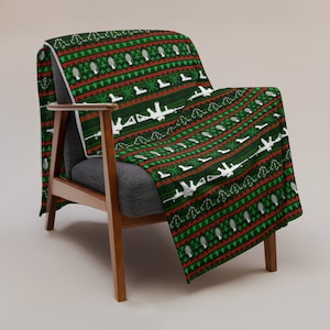 May include: A green and red patterned throw blanket with a white silhouette of a rifle design. The blanket is draped over a wooden armchair.