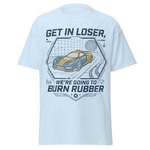 Può includere: Una t-shirt azzurro chiaro con la grafica di un'auto sportiva gialla e blu scuro su una pista. Il testo recita GET IN LOSER, WE'RE GOING TO BURN RUBBER. Il design include una cornice geometrica e una struttura a cupola geodetica.