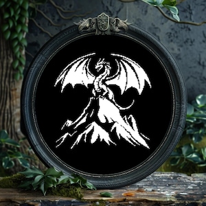 May include: A black and white embroidery design of a dragon perched on a mountain peak. The dragon is facing forward with its wings spread out. The design is framed in a black wooden frame.