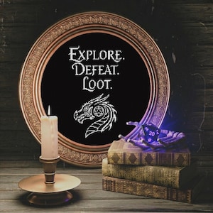 Peut inclure: Une broderie noire encadrée avec les mots "EXPLORE. DEFEAT. LOOT." et un motif de tête de dragon. Une bougie allumée est devant le cadre, avec une pile de livres et un jeu de dés à droite. Le cadre est de couleur bronze.