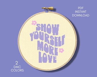 Könnte beinhalten: Ein Kreuzstichmuster mit dem Spruch "Show Yourself More Love" in lila Schrift mit rosa Blumen auf cremefarbenem Hintergrund. Das Muster befindet sich in einem hölzernen Stickrahmen.