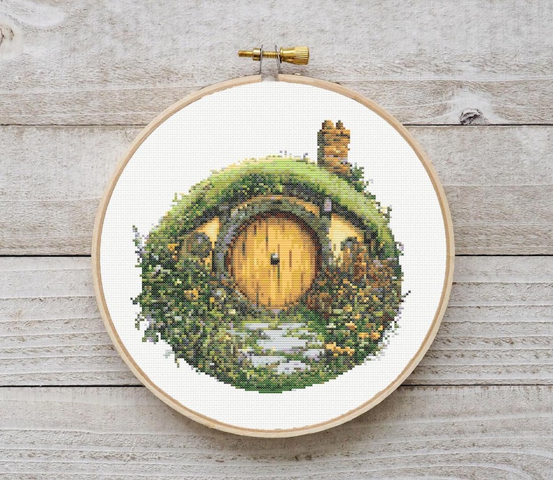 K&ouml;nnte beinhalten: Ein Kreuzstichmuster eines Hobbitlochs mit einer runden, gelben T&uuml;r und gr&uuml;nem Laub.