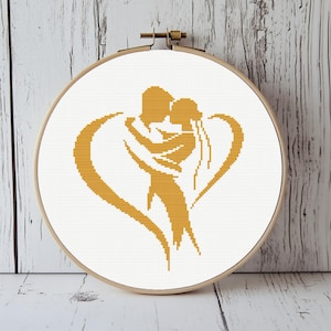 May include: A cross-stitch embroidery hoop featuring a golden silhouette of a couple embracing within a heart shape. The design is set against a white background, framed by a wooden hoop, and mounted on a light wood-grain surface.