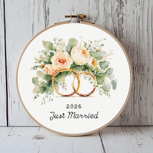 Op de afbeelding: Kruissteekborduurwerk in een houten ring met een bloemenboeket met perzik- en witte rozen, groene bladeren en twee gouden trouwringen. De tekst "2026 Just Married" is eronder geborduurd. De ring staat tegen een lichte houten achtergrond.
