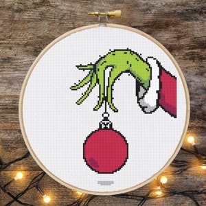 Può includere: Una mano verde con una manica rossa che tiene un ornamento rosso per l'albero di Natale. La mano tiene l'ornamento per una corda.