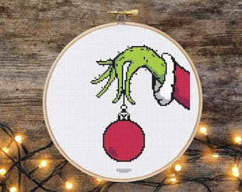 May include: A green hand with a red sleeve holding a red Christmas ornament. The hand is holding the ornament by a string.