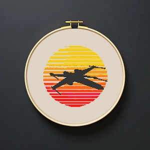 May include: Cross-stitch art featuring a black silhouette of a starfighter against a sunset. The sunset is a gradient of yellow, orange, and red. The artwork is displayed in a round, gold-colored embroidery hoop.