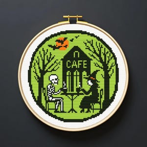 Peut inclure: Broderie au point de croix dans un cercle en bois. Le motif représente un squelette et une sorcière à une table de café, avec le mot "CAFE" au-dessus. Le fond est vert avec des arbres, une lune et des chauves-souris, parfait pour Halloween.