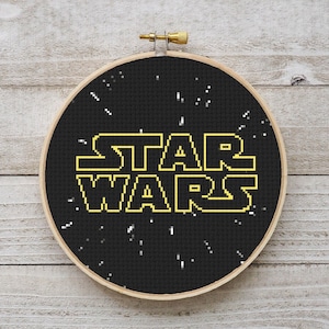 Può includere: Un cerchio da ricamo nero con uno sfondo bianco e le parole "STAR WARS" in lettere gialle pixelate. Lo sfondo è pieno di piccoli punti bianchi.