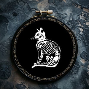 May include: A black cross-stitch embroidery hoop with a white cat skeleton design. The cat's skeleton is visible against the black background. The hoop has a gold-coloured trim. The design is set in a round frame.