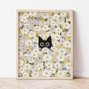 May include: A framed cross-stitch pattern featuring a black cat head with large white eyes peeking out from a dense field of white daisies with yellow centers. The design is rendered in a pixelated style within a light-colored wooden frame.