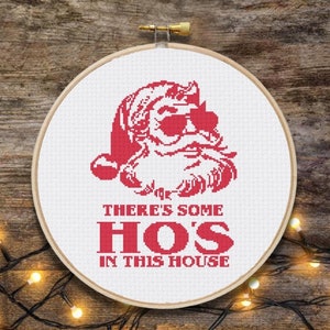 May include: A white embroidery hoop with a red cross-stitch design of Santa Claus wearing sunglasses and the text "There's some HOS in this house".
