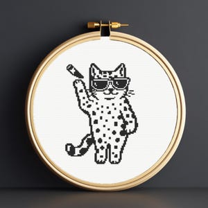 Könnte beinhalten: Kreuzstich-Stickerei einer coolen Katze mit Sonnenbrille und Zigarette. Die Katze hat schwarze Flecken und befindet sich in einem hölzernen Stickrahmen. Das Design ist in Schwarz und Weiß gehalten.