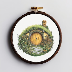 K&ouml;nnte beinhalten: Ein runder Stickrahmen mit wei&szlig;em Hintergrund und einem Kreuzstich-Motiv eines Hobbitlochs mit einer runden gelben T&uuml;r und gr&uuml;nem Laub.