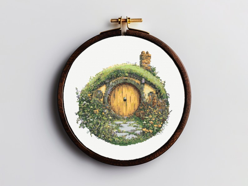 K&ouml;nnte beinhalten: Ein runder Stickrahmen mit wei&szlig;em Hintergrund und einem Kreuzstich-Motiv eines Hobbitlochs mit einer runden gelben T&uuml;r und gr&uuml;nem Laub.
