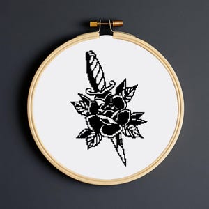 Könnte beinhalten: Schwarzer und weißer Stickrahmen mit einem Design aus einer schwarzen Rose und einem Dolch.