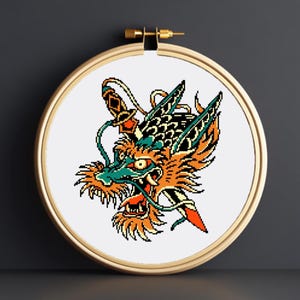 Peut inclure: Un motif de broderie au point de croix représentant une tête de dragon dans un cercle à broder en bois. Le dragon est orange, vert et noir, avec une épée. L'ensemble est présenté sur un fond gris foncé.