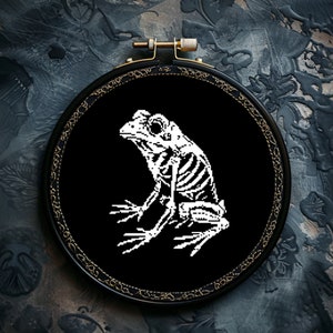 May include: A black embroidery hoop featuring a white skeletal frog design. The frog is sitting on a black background.