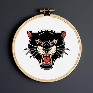 Op de afbeelding: Een zwart panterhoofd kruissteekontwerp in een houten borduurring. De panter heeft rode ogen en een open mond, die een rode tong onthult. Het ontwerp staat op een witte achtergrond, met een donkergrijze achtergrond.