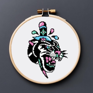 May include: A cross-stitch embroidery hoop featuring a black panther head with a dagger design. The panther has pink, teal, and white accents, with a pixelated aesthetic. The hoop is made of wood and has a brass screw.