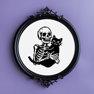 May include: Black and white embroidery hoop with a skeleton holding a black cat. The skeleton is sitting down and the cat is curled up around it. The embroidery is in a black frame.