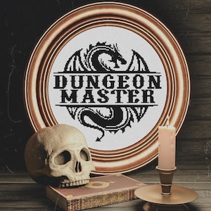 Op de afbeelding: Een ronde koperkleurige lijst met een zwart kruissteekontwerp van een draak-silhouet met de tekst "DUNGEON MASTER" in het midden. Een beige menselijke schedel, een rechthoekig boek en een brandende kaars staan aan de voorzijde.