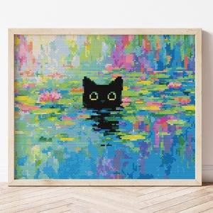 May include: A framed cross-stitch art piece featuring a black cat with green eyes floating in water. The water is a mosaic of blues, greens, pinks, and yellows, creating a vibrant, colorful scene. The frame is a light wood color.