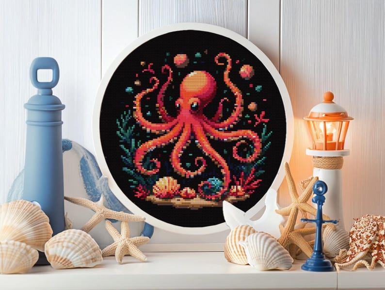 May include: A round embroidery hoop with a black background and a colorful octopus with eight tentacles surrounded by coral, seashells, and bubbles. The octopus is red with a white belly and blue eyes.