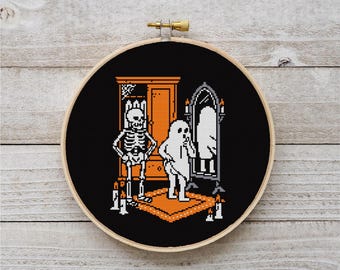 May include: A Halloween cross-stitch design in a wooden hoop. The image shows a skeleton, a ghost, a mirror, and a wardrobe, all in a pixelated style. The colour scheme includes black, white, orange, and beige.