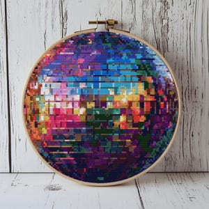 Può includere: Un colorato disegno a punto croce in un telaio da ricamo in legno. L'opera presenta un mosaico di colori vivaci, tra cui rosa, blu, viola e giallo. Il telaio in legno è fissato con un fermaglio in ottone.